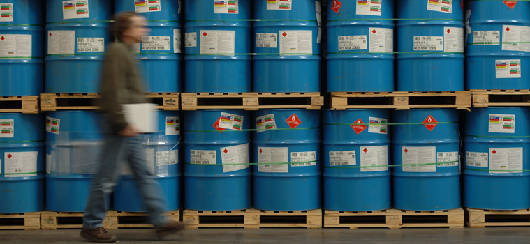 A male worker walking past labelled chemical drums holding a clipboard 