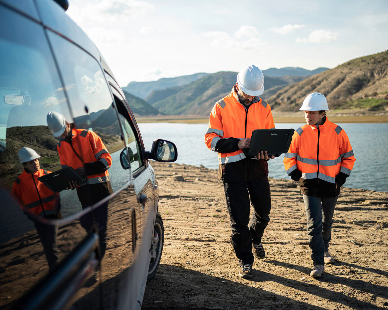 Two workers in high-visibility orange jackets and white hard hats walk beside a vehicle on a dirt path near a lake, with mountains in the background. One worker holds a tablet, and the other walks alongside.